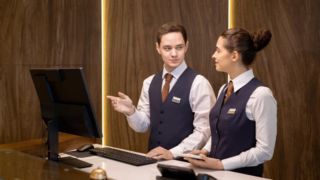 Young hotel receptionists looking at colleague with touchpad and pointing at computer screen